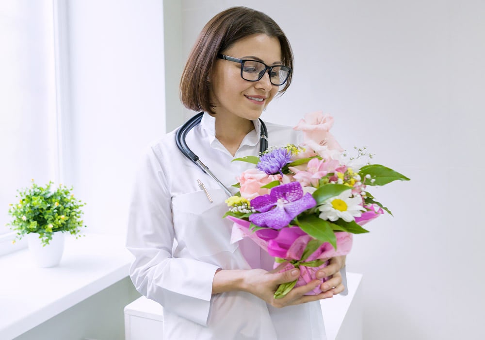 A woman in a white coat, with a stethoscope around her neck, holds a bouquet of colorful flowers in a bright, white room with a potted plant beside a window.