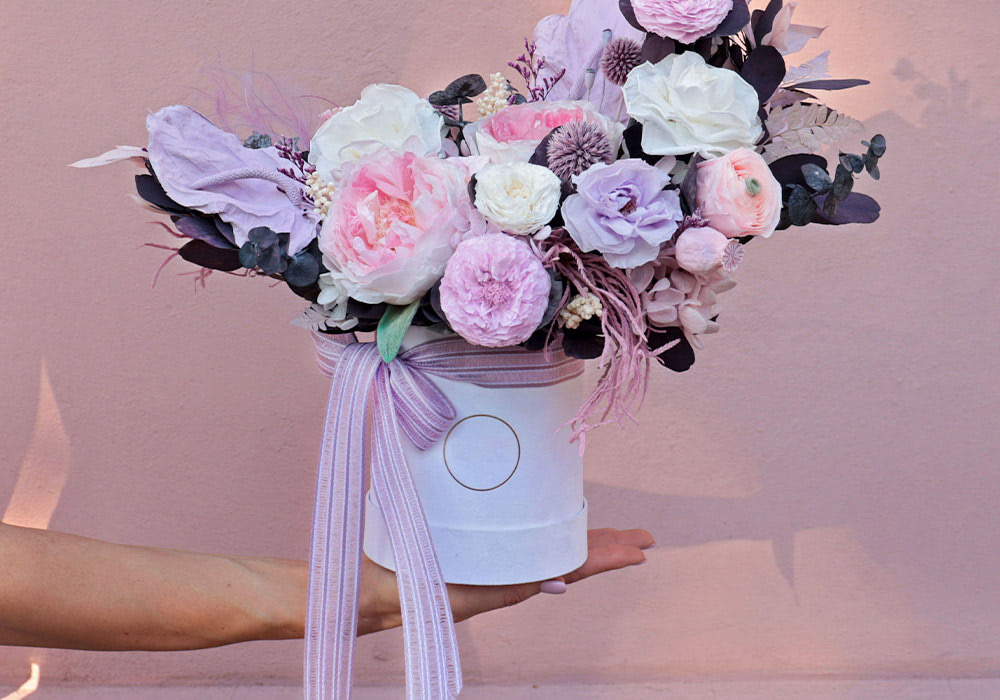 A hand holds a white cylindrical vase filled with pink, white, and lavender flowers, adorned with a striped ribbon, against a pale pink background.